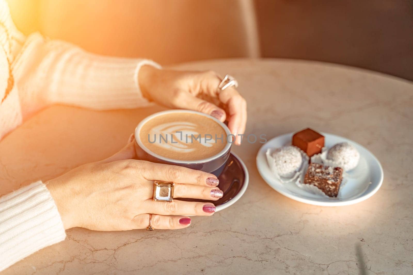 A cup of fresh cappuccino coffee in the hands of a woman on a fashionable background of a white marble table, next to a plate with sweets. Coffee addiction.