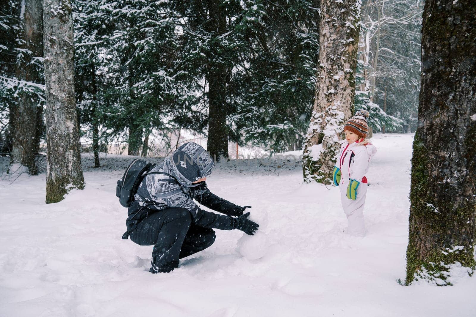 Little girl stands in a snowy forest and looks at her mother making a snowman while squatting. High quality photo