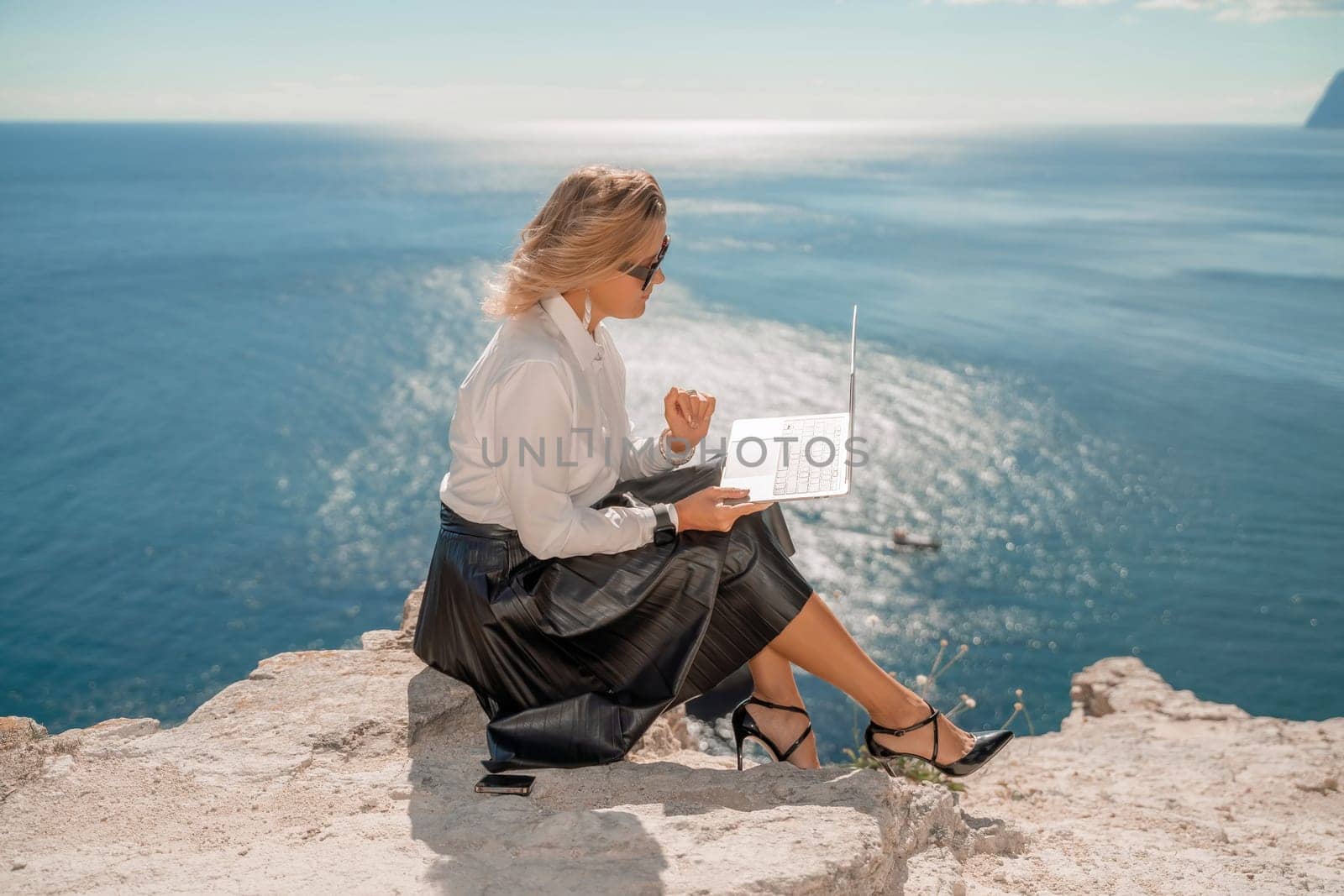 Business woman on nature in white shirt and black skirt. She works with an iPad in the open air with a beautiful view of the sea. The concept of remote work