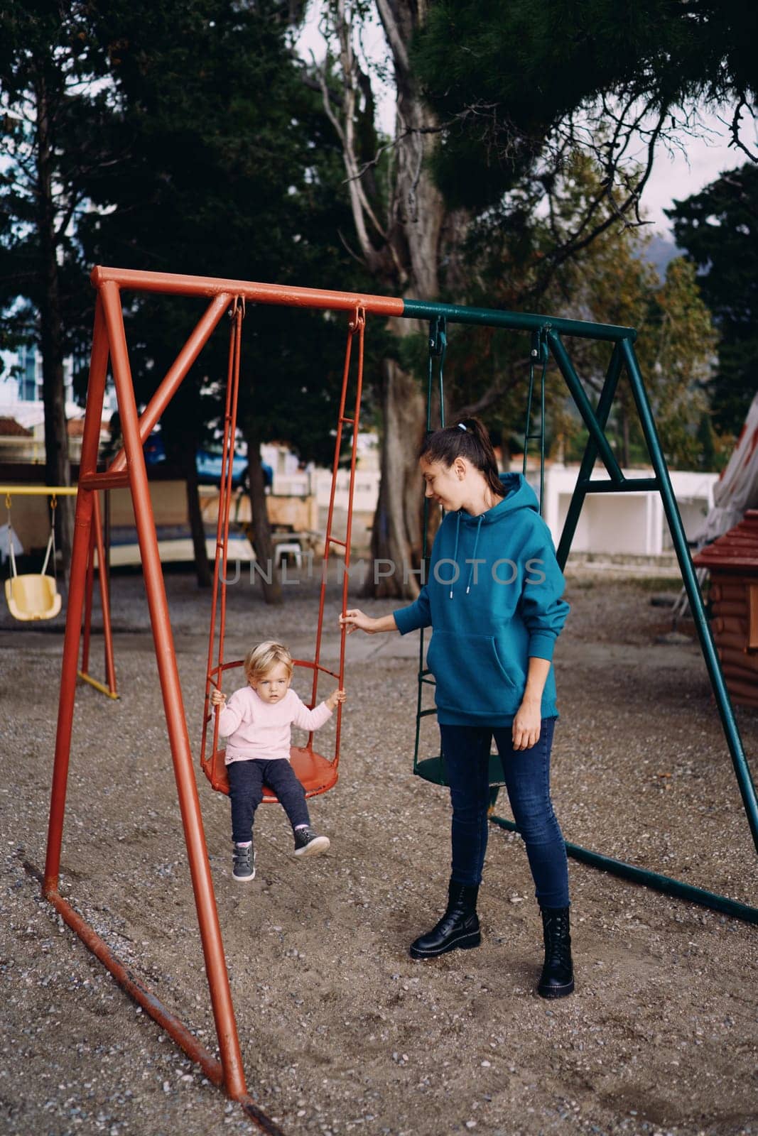 Mom stands next to the swing on the playground, with a little girl sitting on it by Nadtochiy