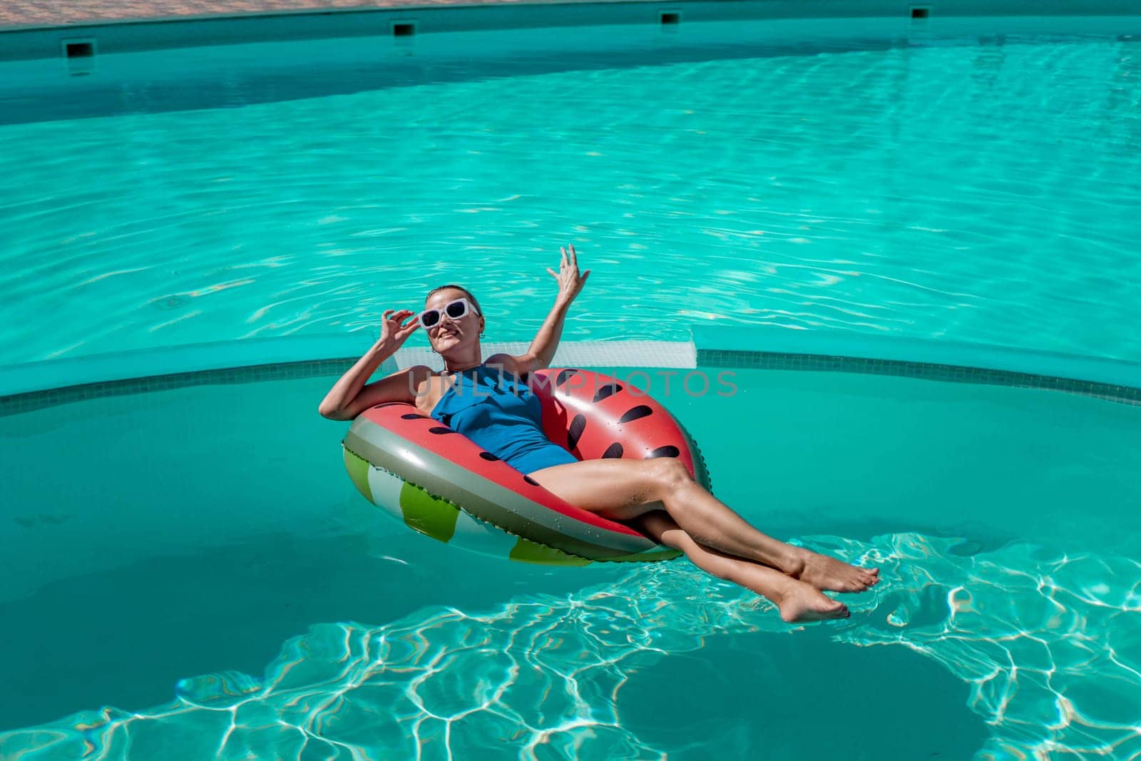 Happy woman in a swimsuit and sunglasses floating on an inflatable ring in the form of a watermelon, in the pool during summer holidays and vacations. Summer concept