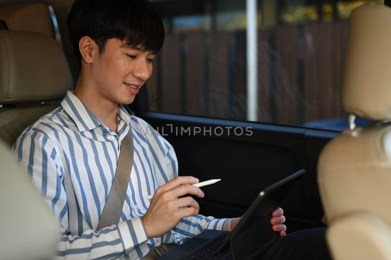 Smiling young businessman working on digital tablet while sitting on the back seat of the car. by prathanchorruangsak