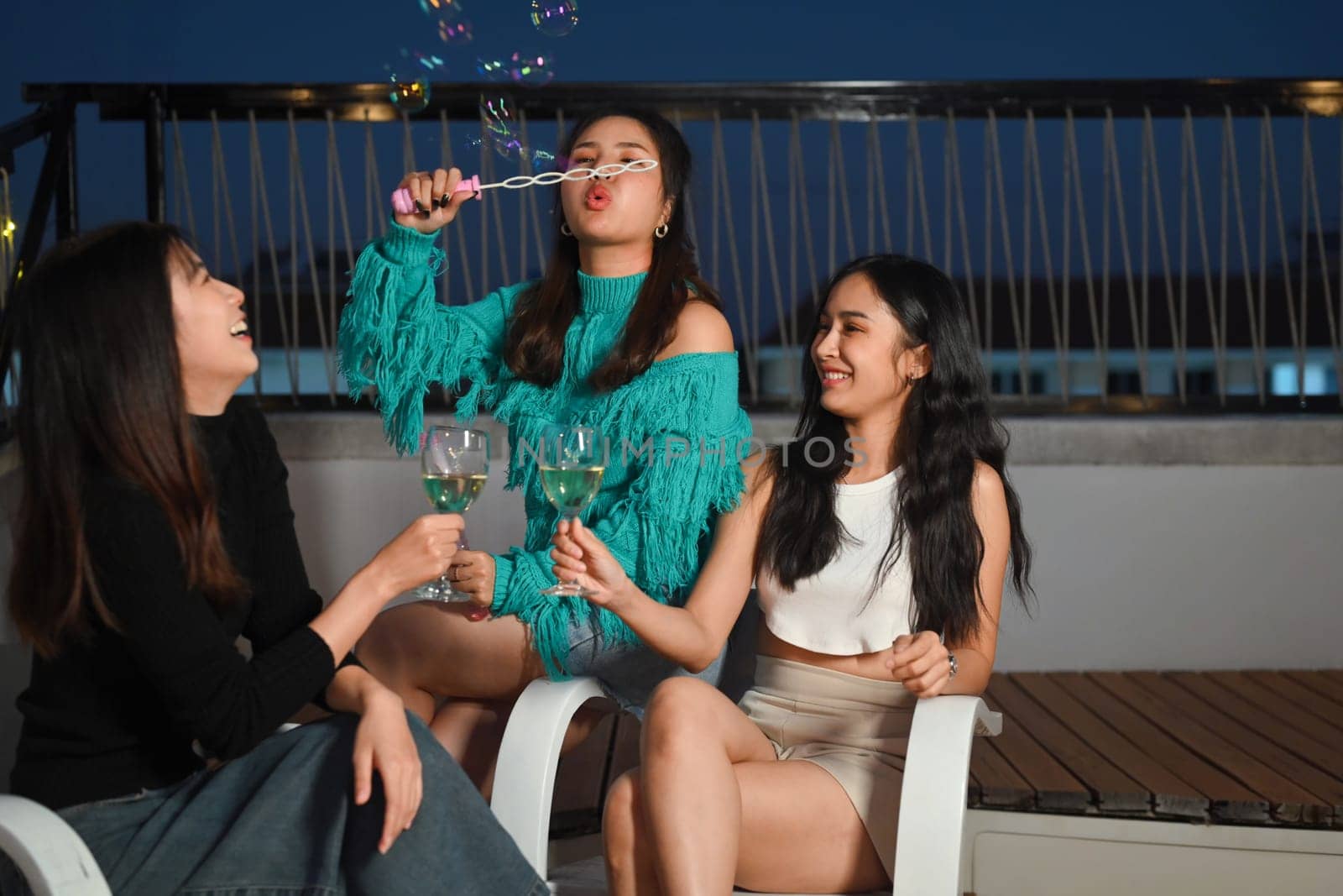 Happy young women friends blowing soap bubbles, having fun toasting wine glasses on a rooftop party