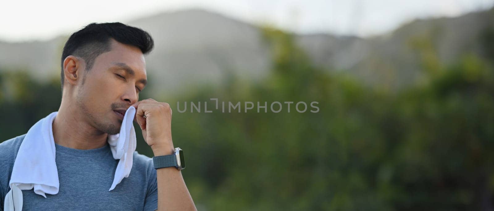 Young athletic man wiping sweat from his forehead during workout outdoors by prathanchorruangsak