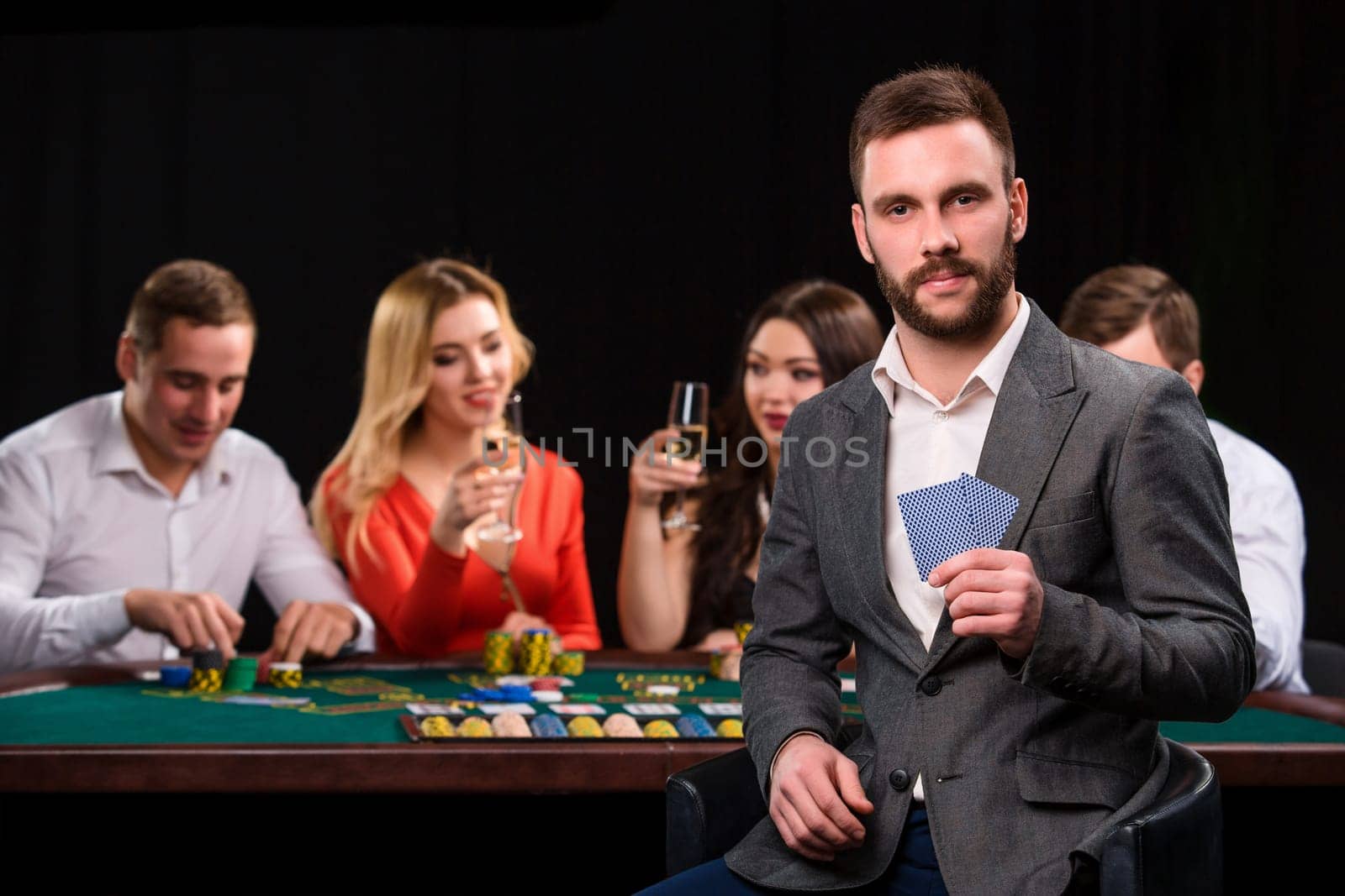 Poker players in casino with cards and chips on black background. A handsome man in the foreground, behind a game table.