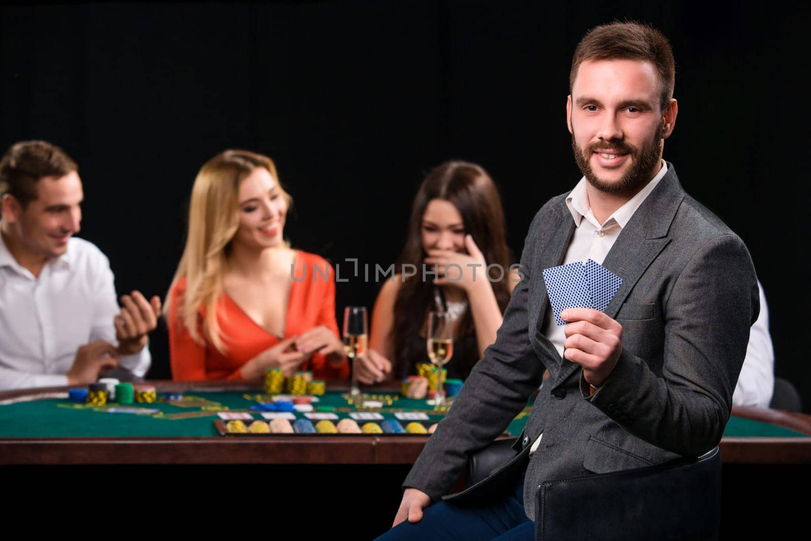 Poker players in casino with cards and chips on black background. A handsome man in the foreground, behind a game table.