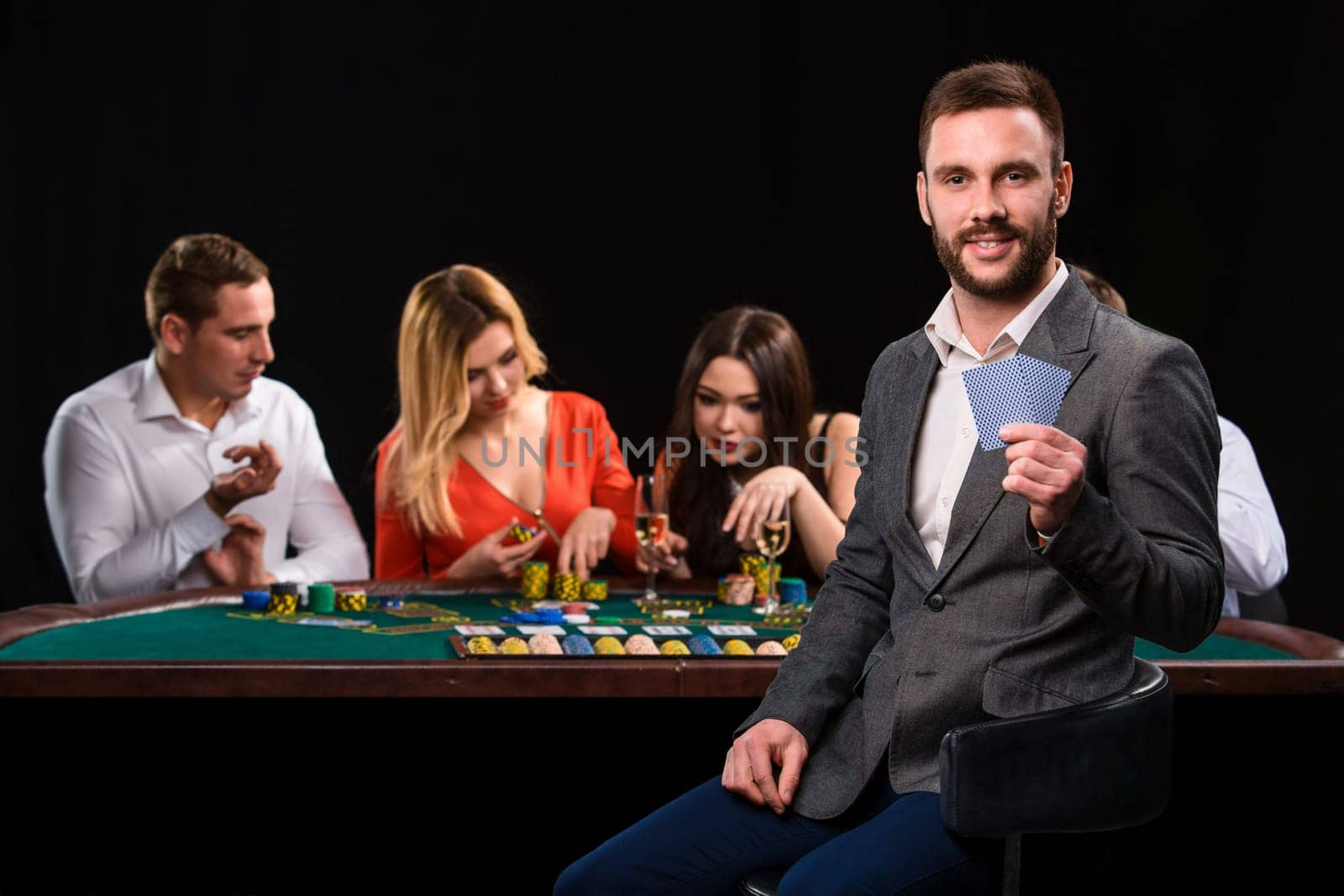 Poker players in casino with cards and chips on black background. A handsome man in the foreground, behind a game table.