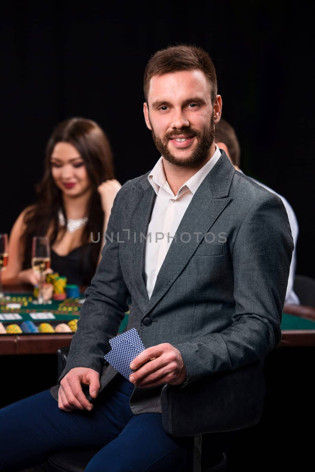Poker players in casino with cards and chips on black background. A handsome man in the foreground, behind a game table.