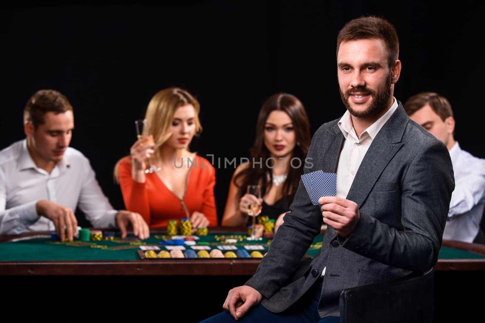 Poker players in casino with cards and chips on black background. A handsome man in the foreground, behind a game table.