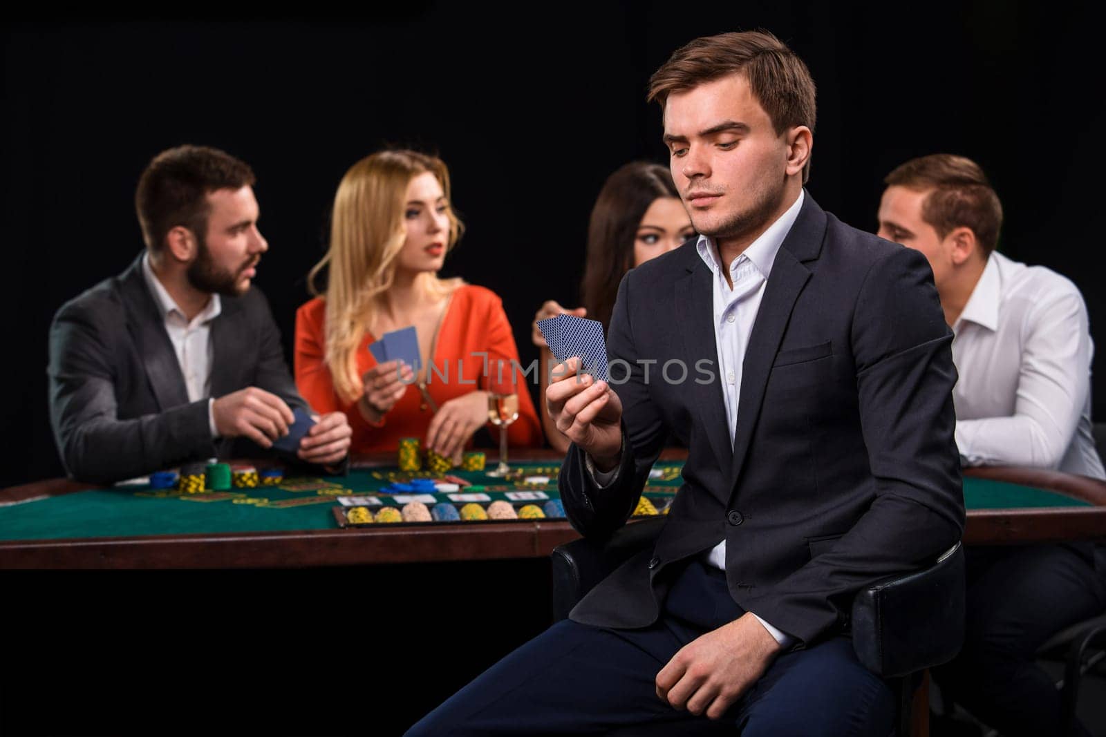 Young people playing poker at the table. Handsome man with cards in hand sitting in the foreground. Casino