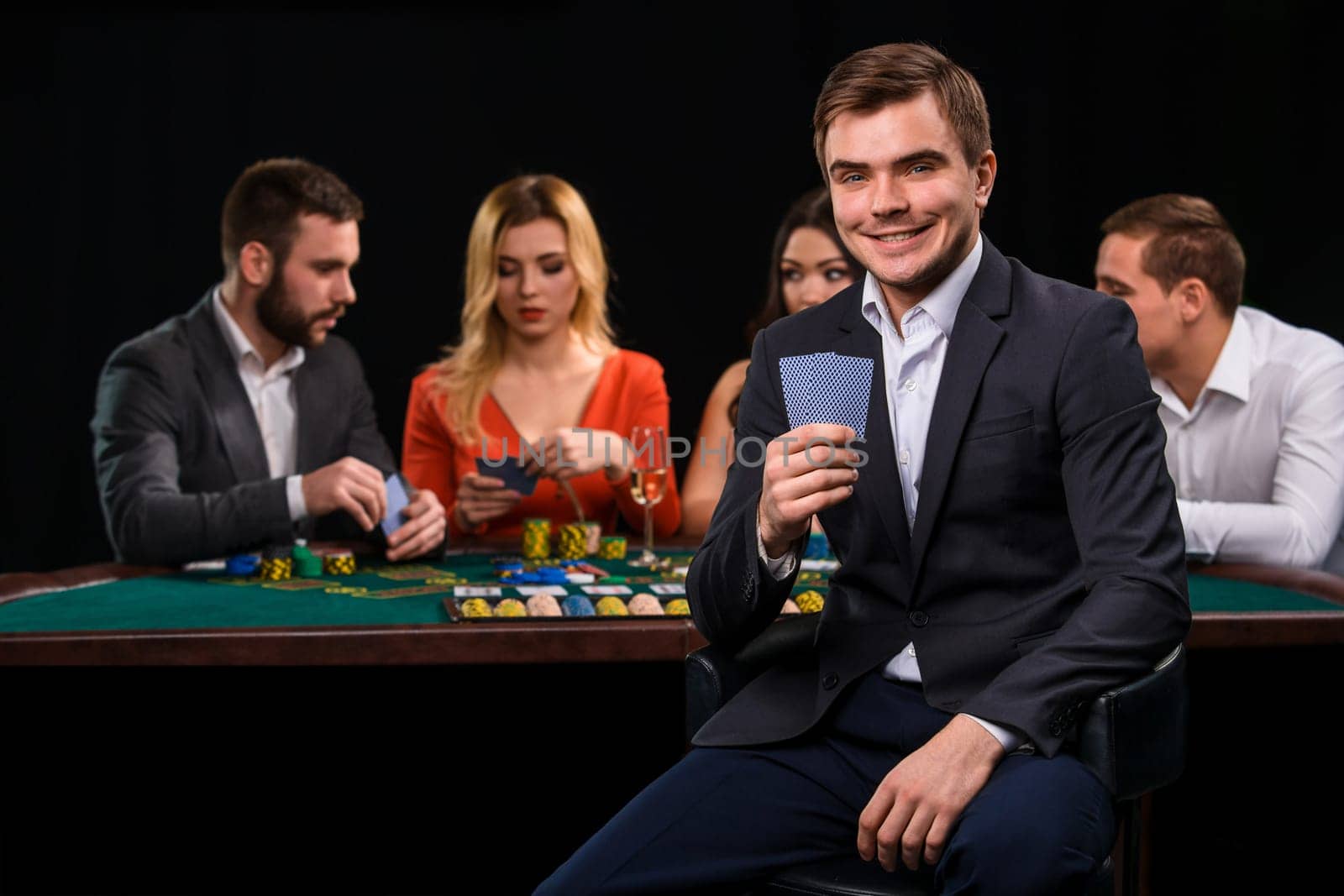Young people playing poker at the table. Handsome man with cards in hand sitting in the foreground. Casino