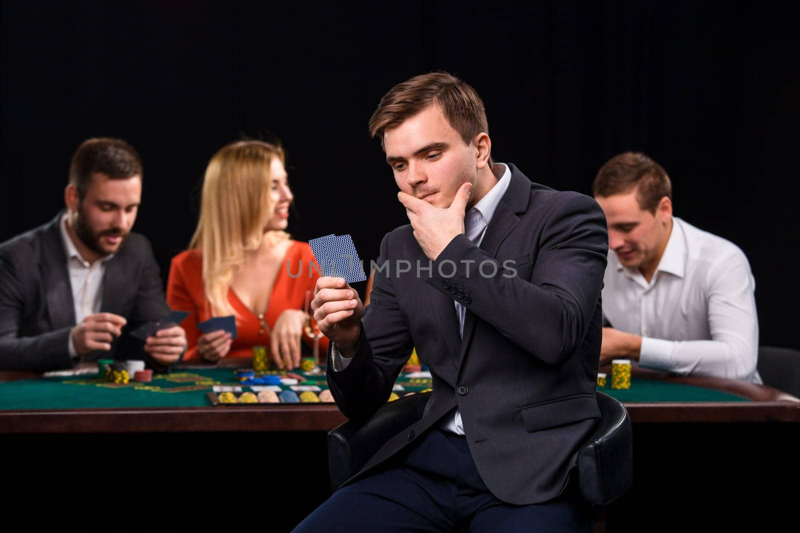 Young people playing poker at the table. Handsome man with cards in hand sitting in the foreground. Casino