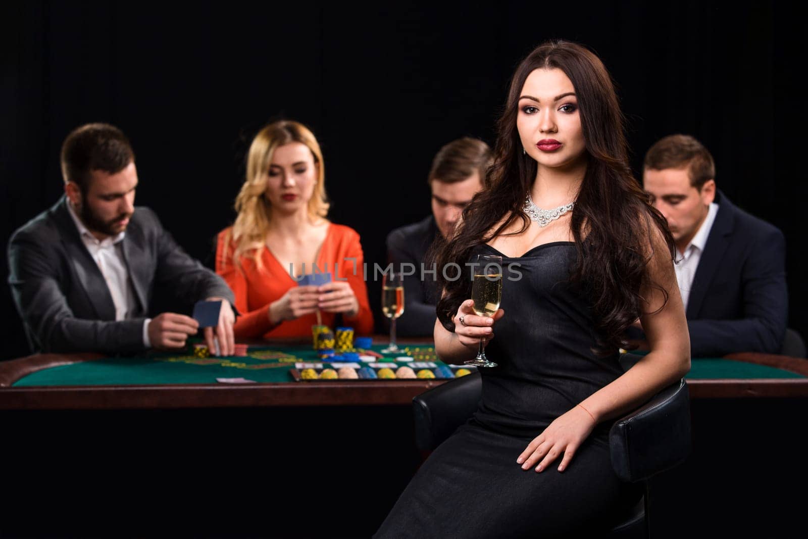 Young people playing poker at the table. Luxury women in dresses with a glass of champagne in hand sitting in the foreground. Casino
