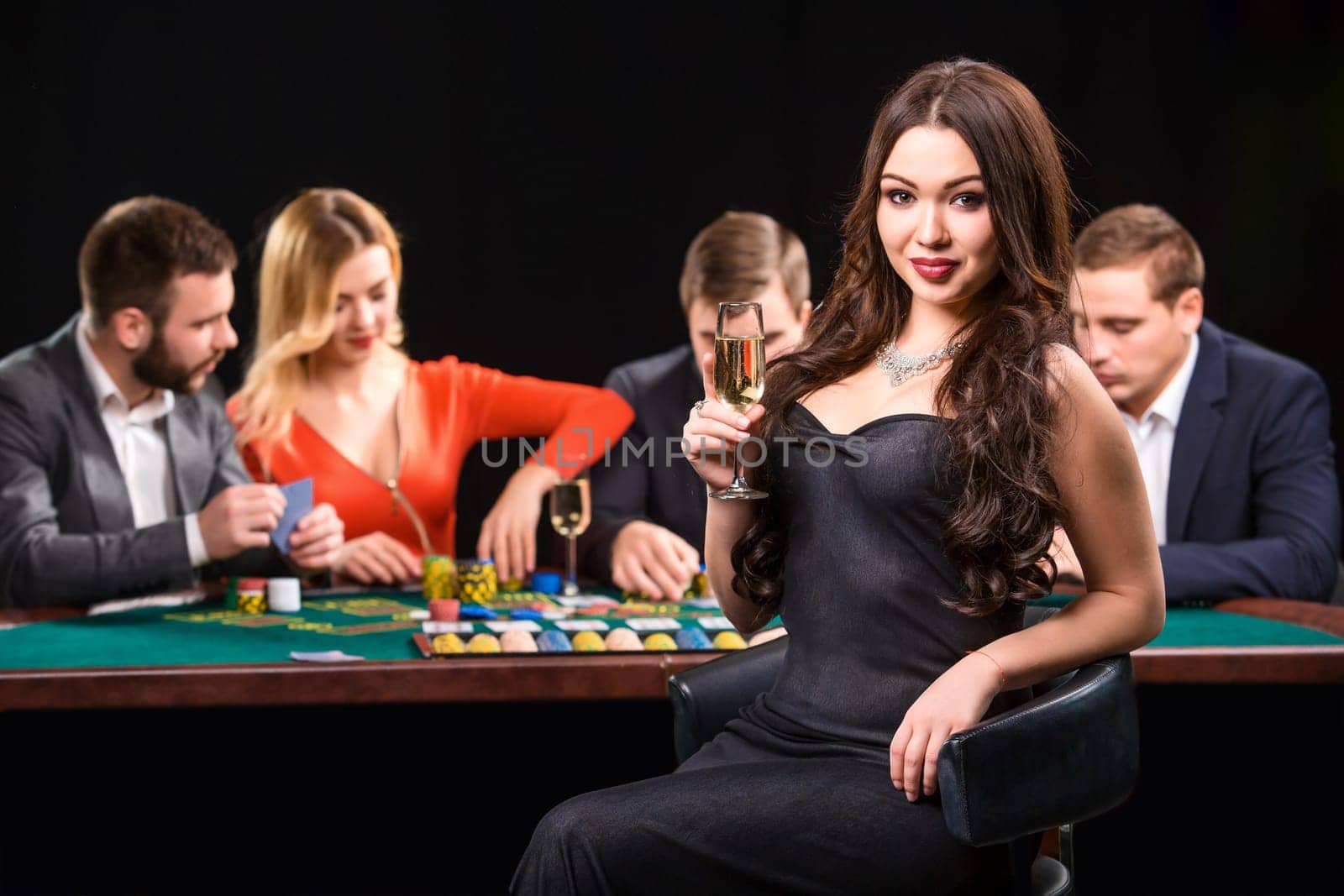 Young people playing poker at the table. Luxury women in dresses with a glass of champagne in hand sitting in the foreground. Casino