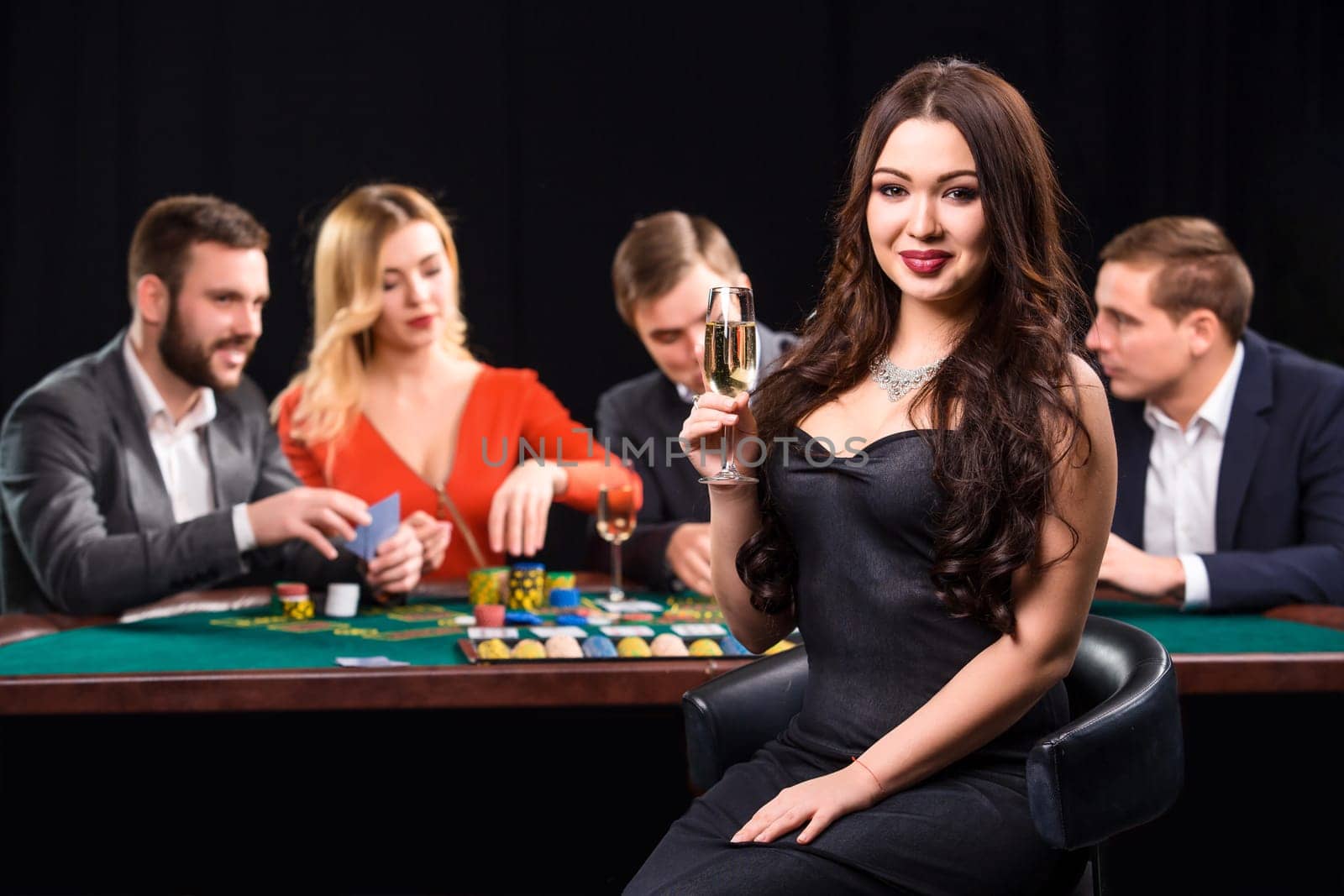 Young people playing poker at the table. Luxury women in dresses with a glass of champagne in hand sitting in the foreground. Casino