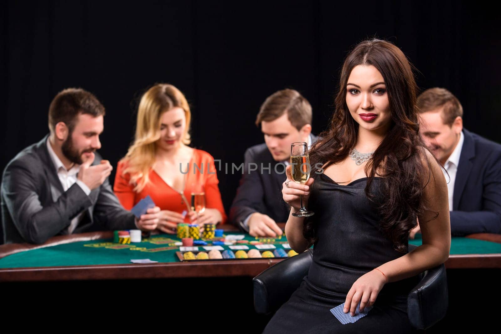 Young people playing poker at the table. Luxury women in dresses with a glass of champagne in hand sitting in the foreground. Casino