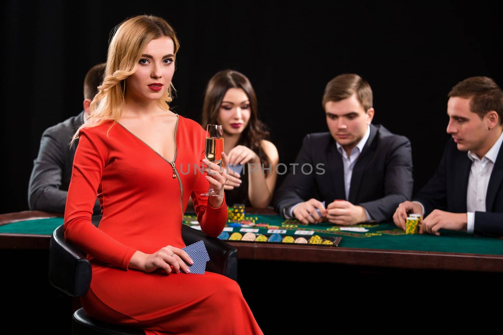 Young people playing poker at the table. Luxury women in dresses with a glass of champagne in hand sitting in the foreground. Casino