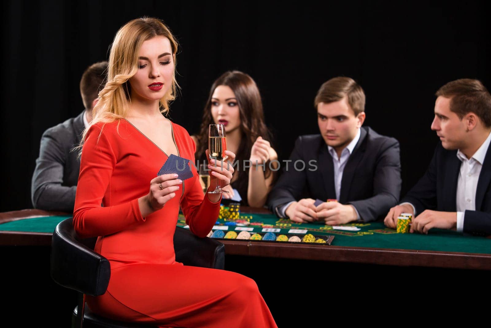 Young people playing poker at the table. Luxury women in dresses with a glass of champagne in hand sitting in the foreground. Casino