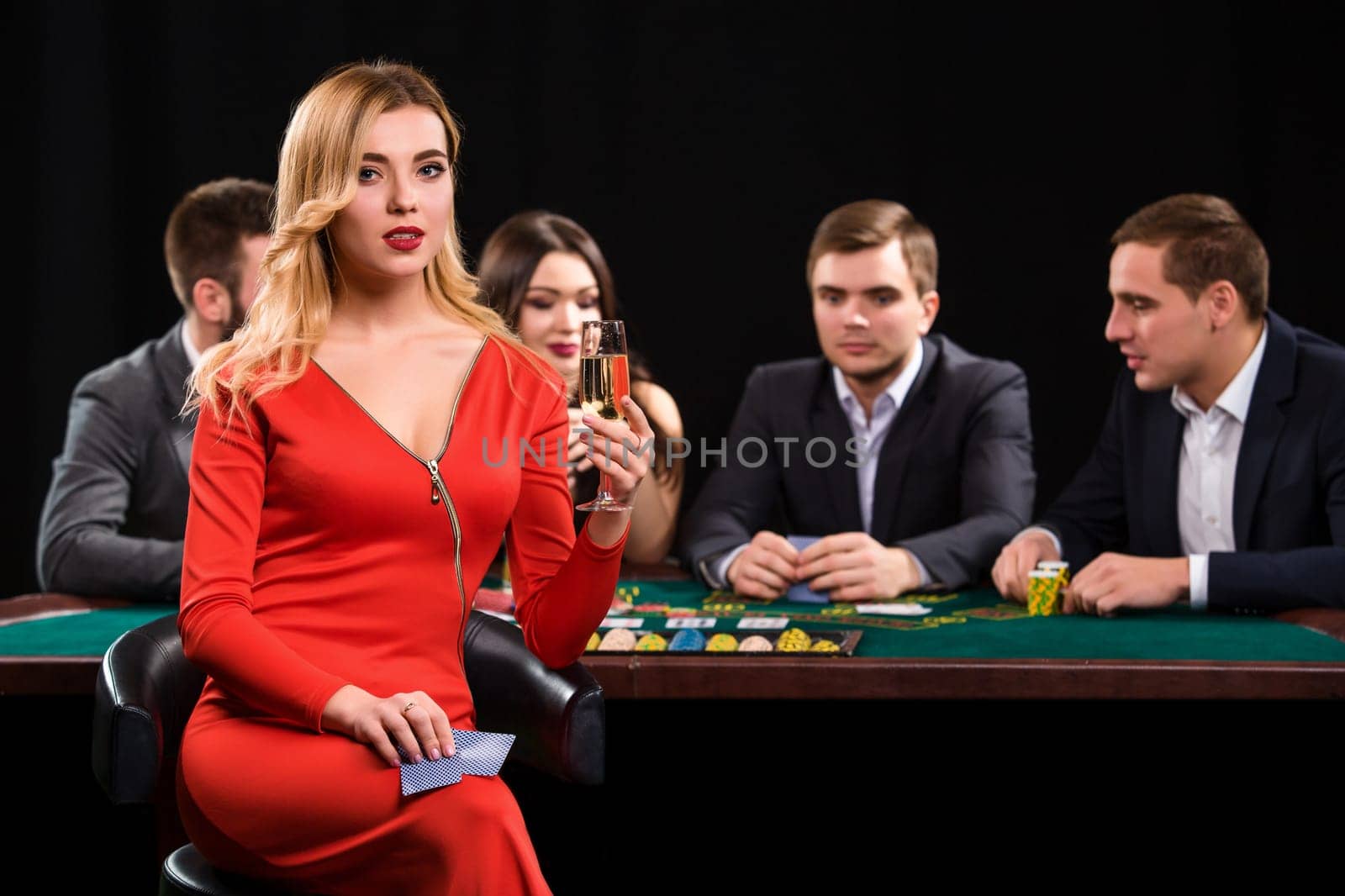 Young people playing poker at the table. Luxury women in dresses with a glass of champagne in hand sitting in the foreground. Casino