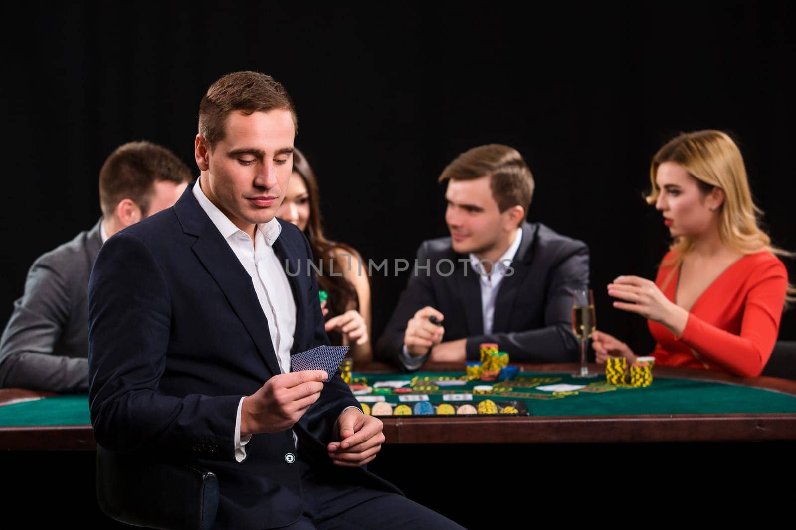 Young people playing poker at the table. Handsome man with cards in hand sitting in the foreground. Casino