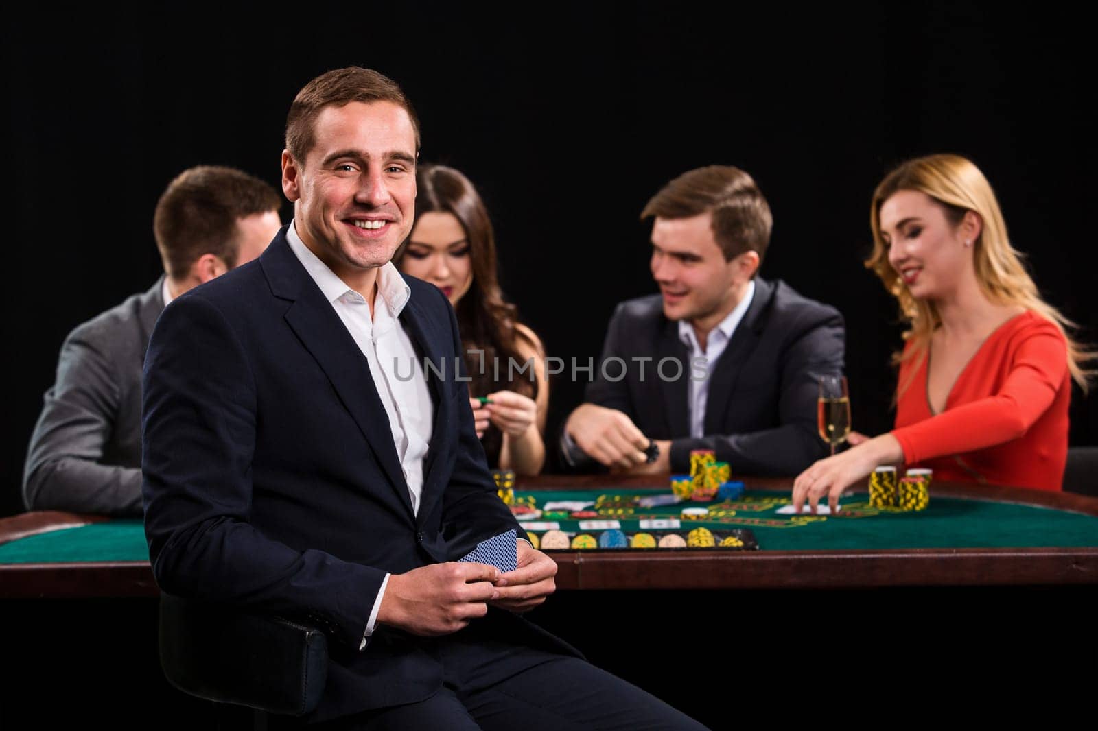 Young people playing poker at the table. Handsome man with cards in hand sitting in the foreground. Casino