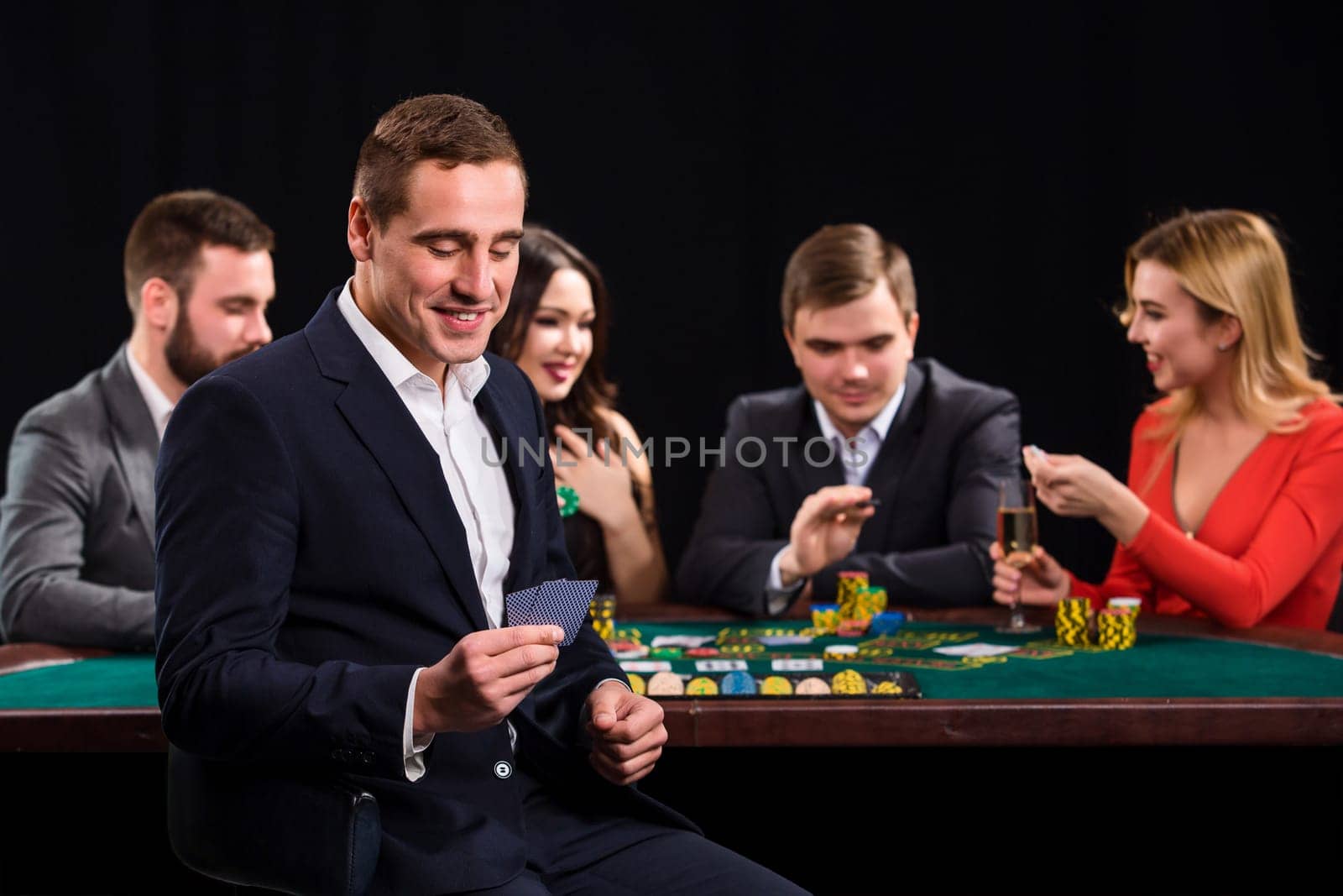 Young people playing poker at the table. Handsome man with cards in hand sitting in the foreground. Casino