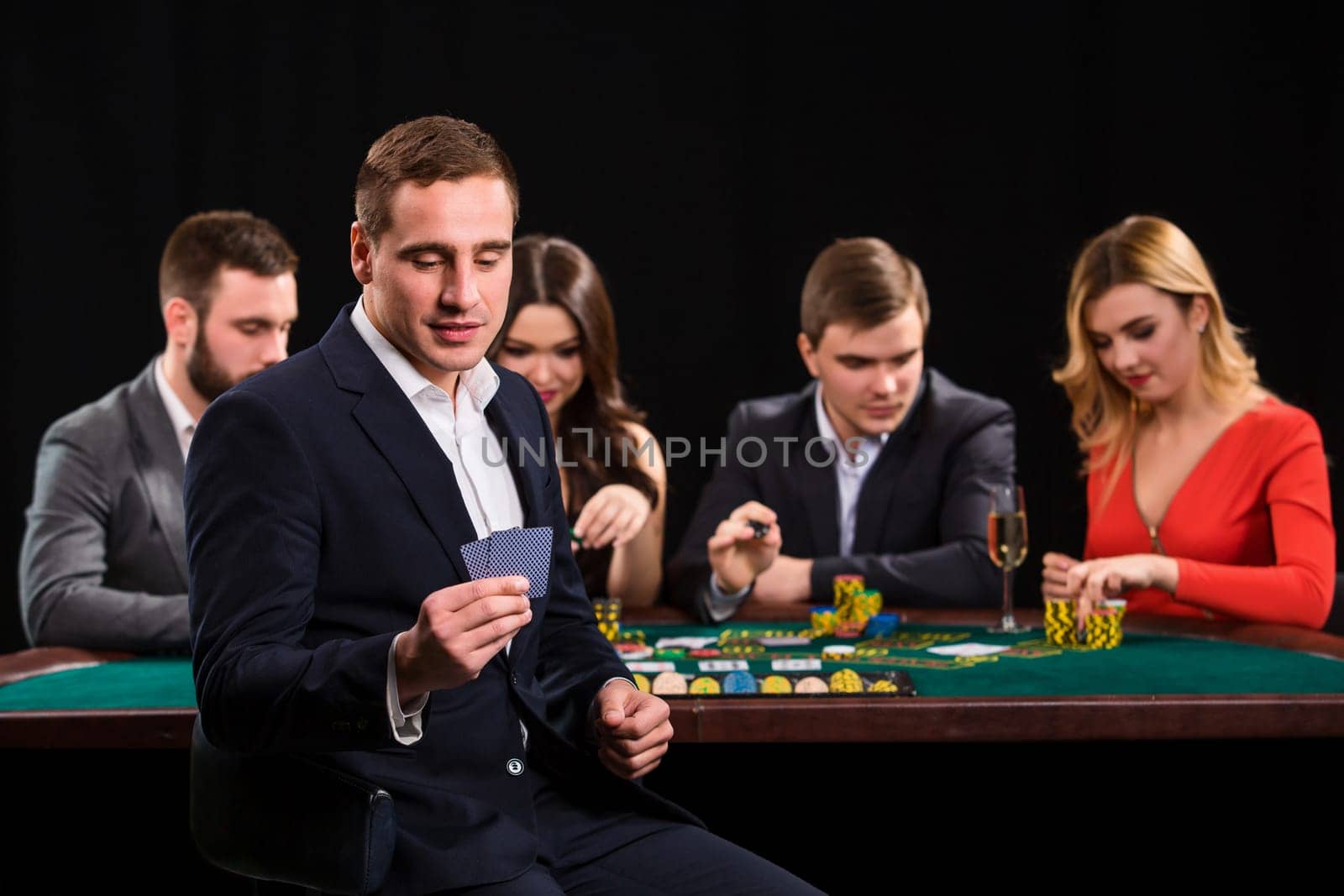 Young people playing poker at the table. Handsome man with cards in hand sitting in the foreground. Casino