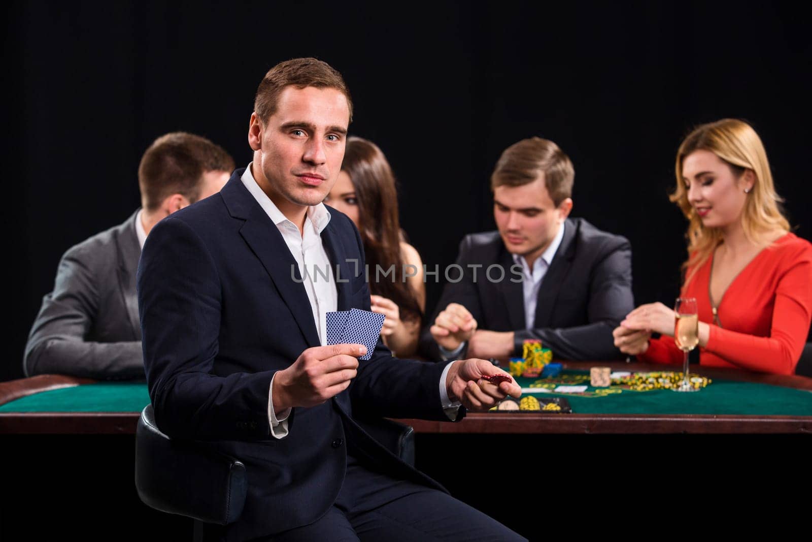 Poker players in casino with cards and chips on black background. A handsome man in the foreground, behind a game table.