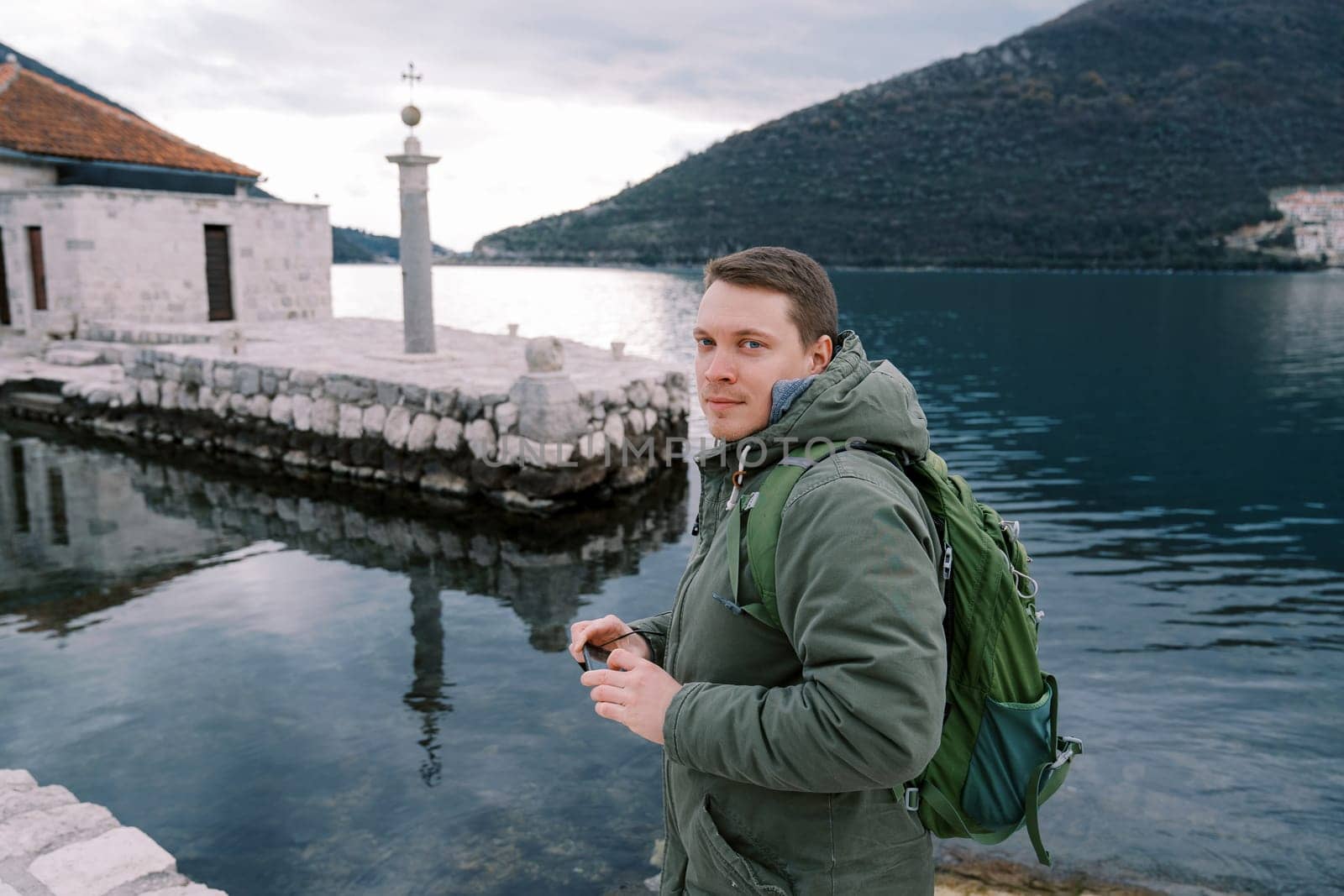 Young man with a smartphone in his hands stands on a stone pier near an ancient building by Nadtochiy