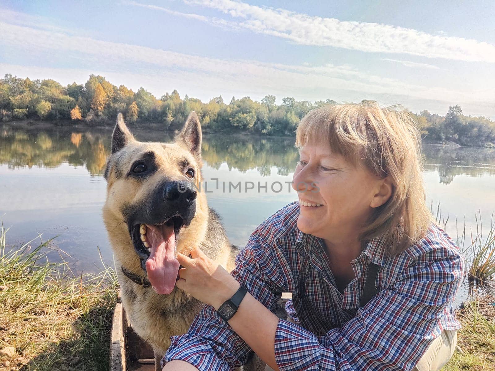 Adult girl with shepherd dog taking selfie near water of river or lake. Middle aged woman and big pet on nature. Friendship, love, fun, hugs by keleny