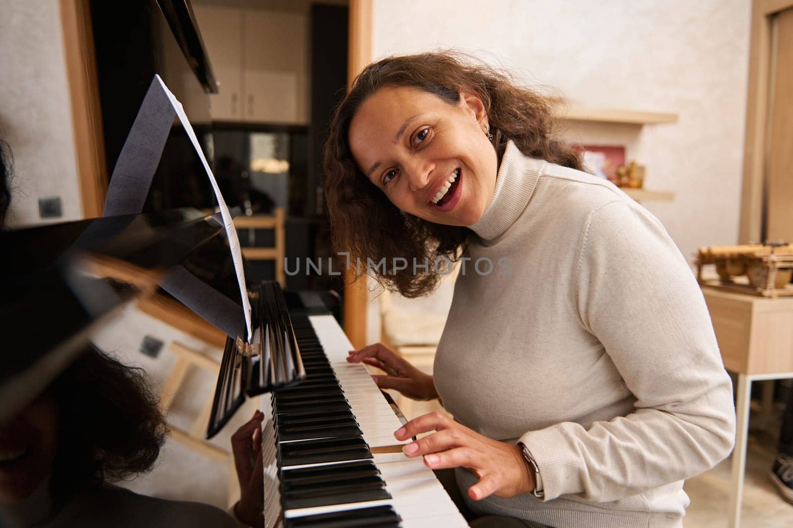 Joyful African American young pretty woman in casual clothes, playing jazz melody on piano at home, smiling broadly looking at the camera