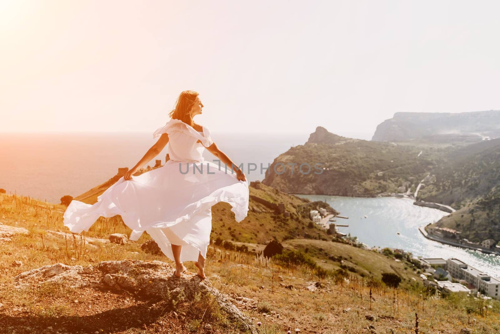 Happy woman in a white dress and hat stands on a rocky cliff above the sea, with the beautiful silhouette of hills in thick fog in the background