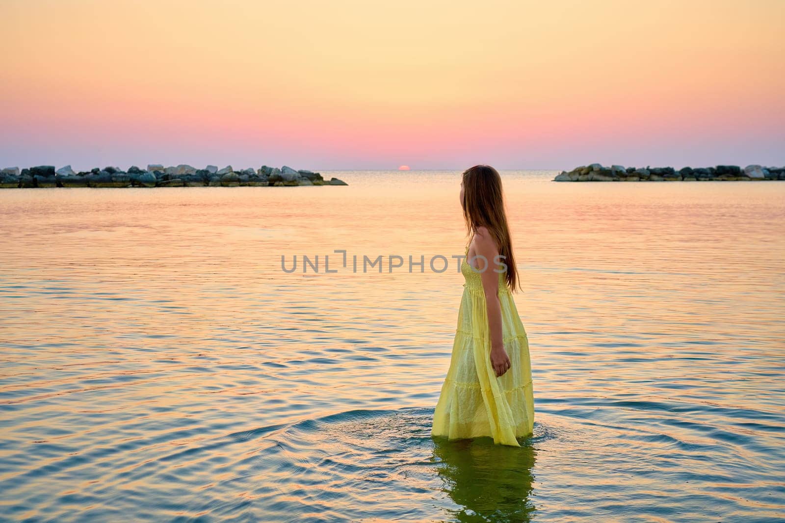 A young woman in a bright yellow dress meets the sunrise, joyfully runs through the sea water