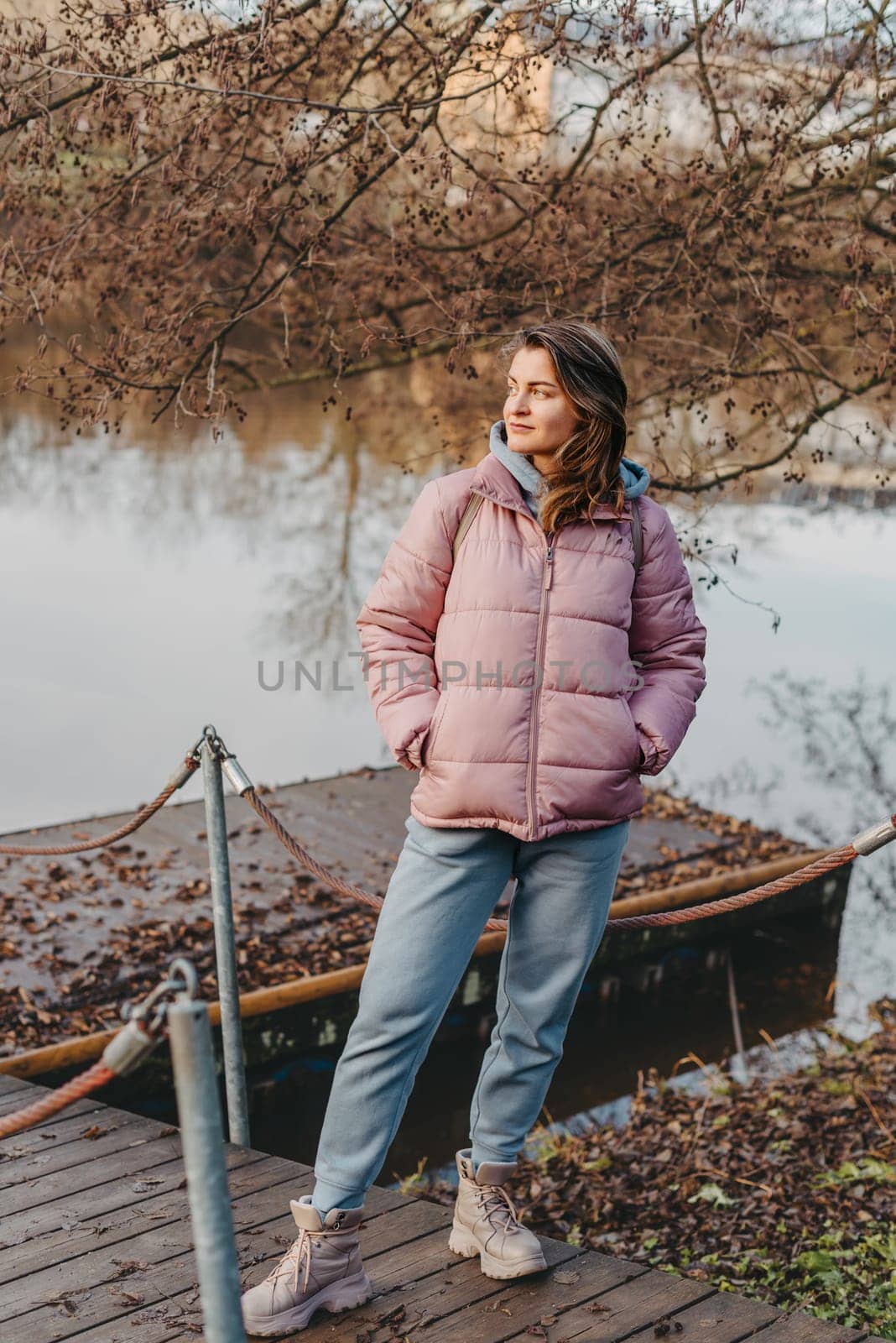 A young woman standing at the shore looking at the river in autumn sunny day. Street view, copy space for text, travel photo. Happy tourist woman on the bank of the river in autumn in warm clothes. Tourists enjoy their vacation, winter season. Romantic look and travel concept. A joyful mood in a Caucasian girl. Winter Wonderland: Enchanting Girl by the Riverside in Autumn.