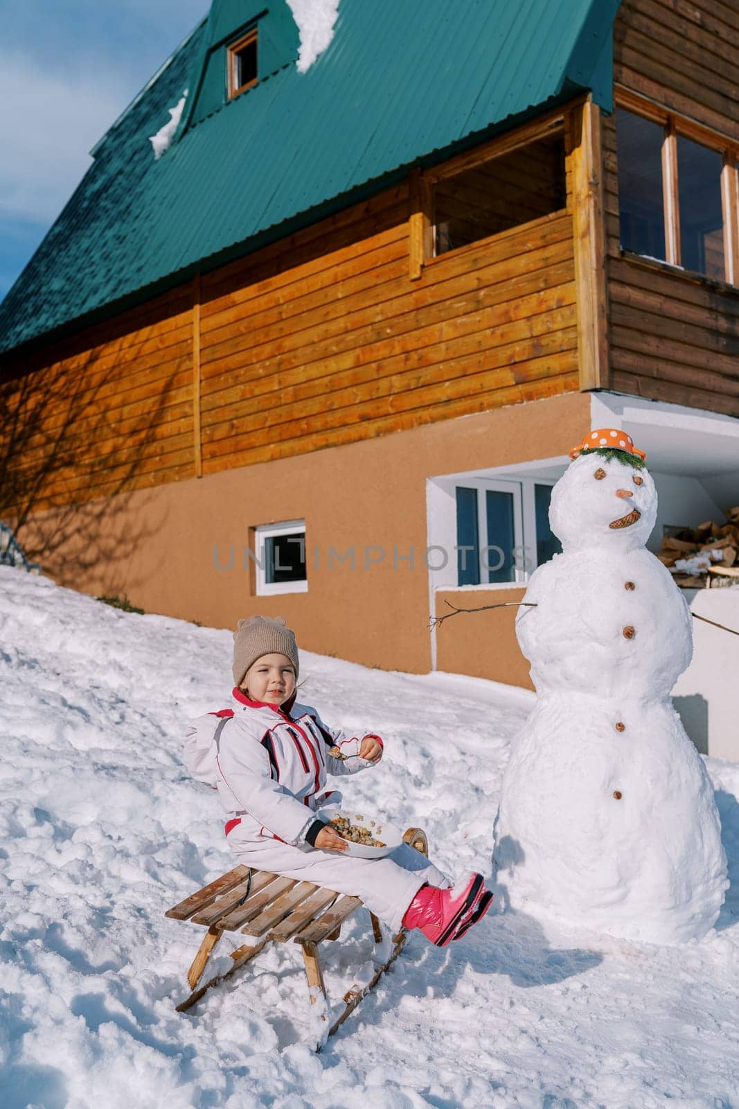 Little girl eats porridge from a plate, sitting on a sleigh near a snowman near the house by Nadtochiy
