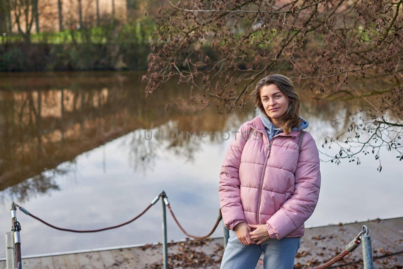 A young woman standing at the shore looking at the river in autumn sunny day. Street view, copy space for text, travel photo. Happy tourist woman on the bank of the river in autumn in warm clothes. Tourists enjoy their vacation, winter season. Romantic look and travel concept. A joyful mood in a Caucasian girl. Winter Wonderland: Enchanting Girl by the Riverside in Autumn.