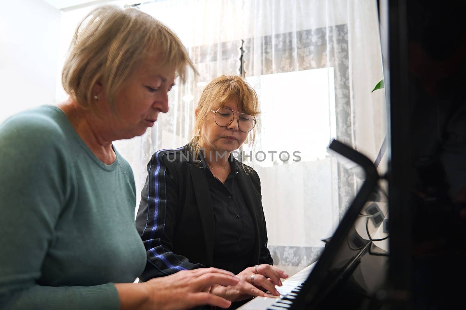A mature Caucasian woman teaches her friend to play the piano. Music lesson by artgf