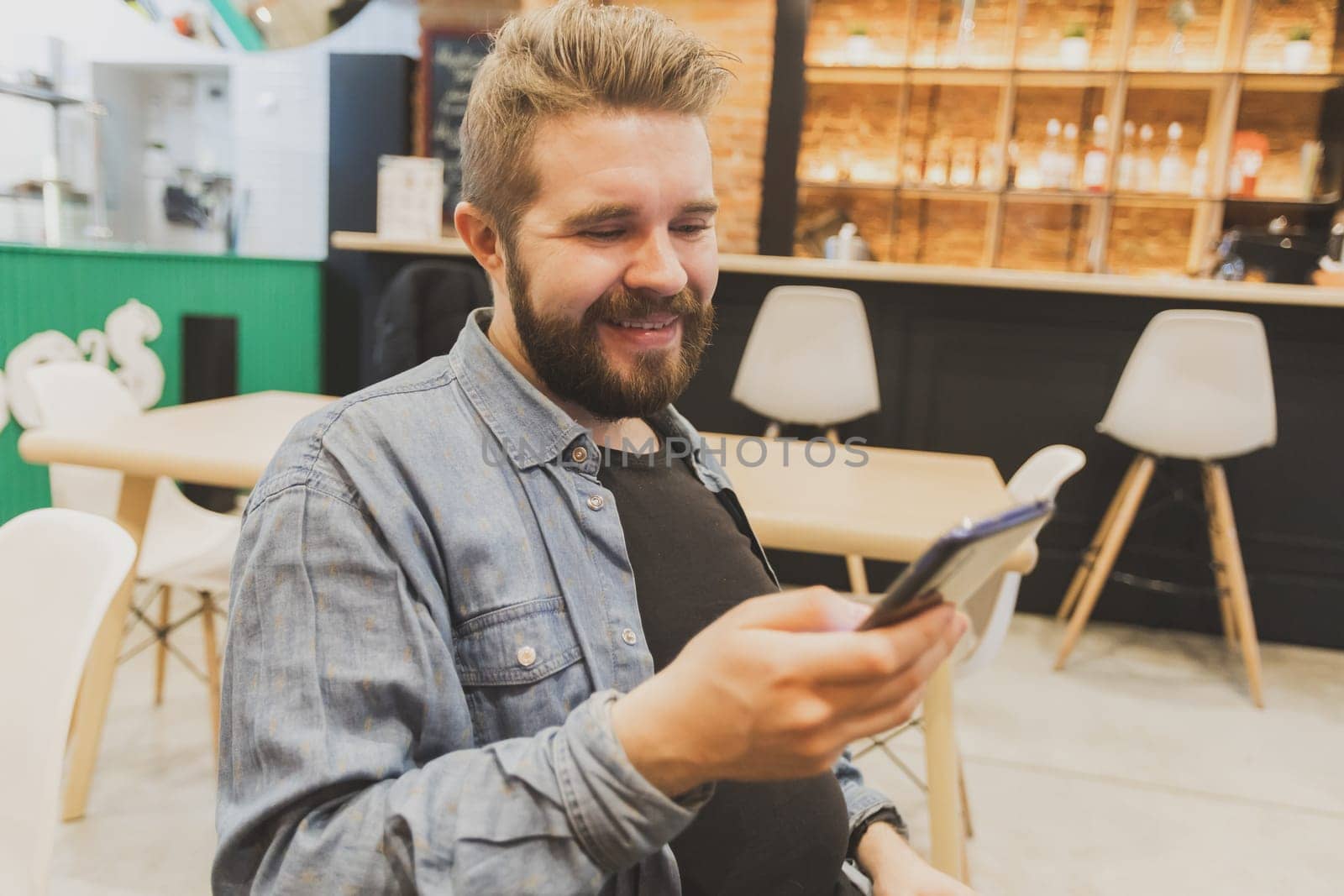 Bearded male using smartphone at modern coffee shop and chatting online messaging on mobile phone. Social networks and reading blog