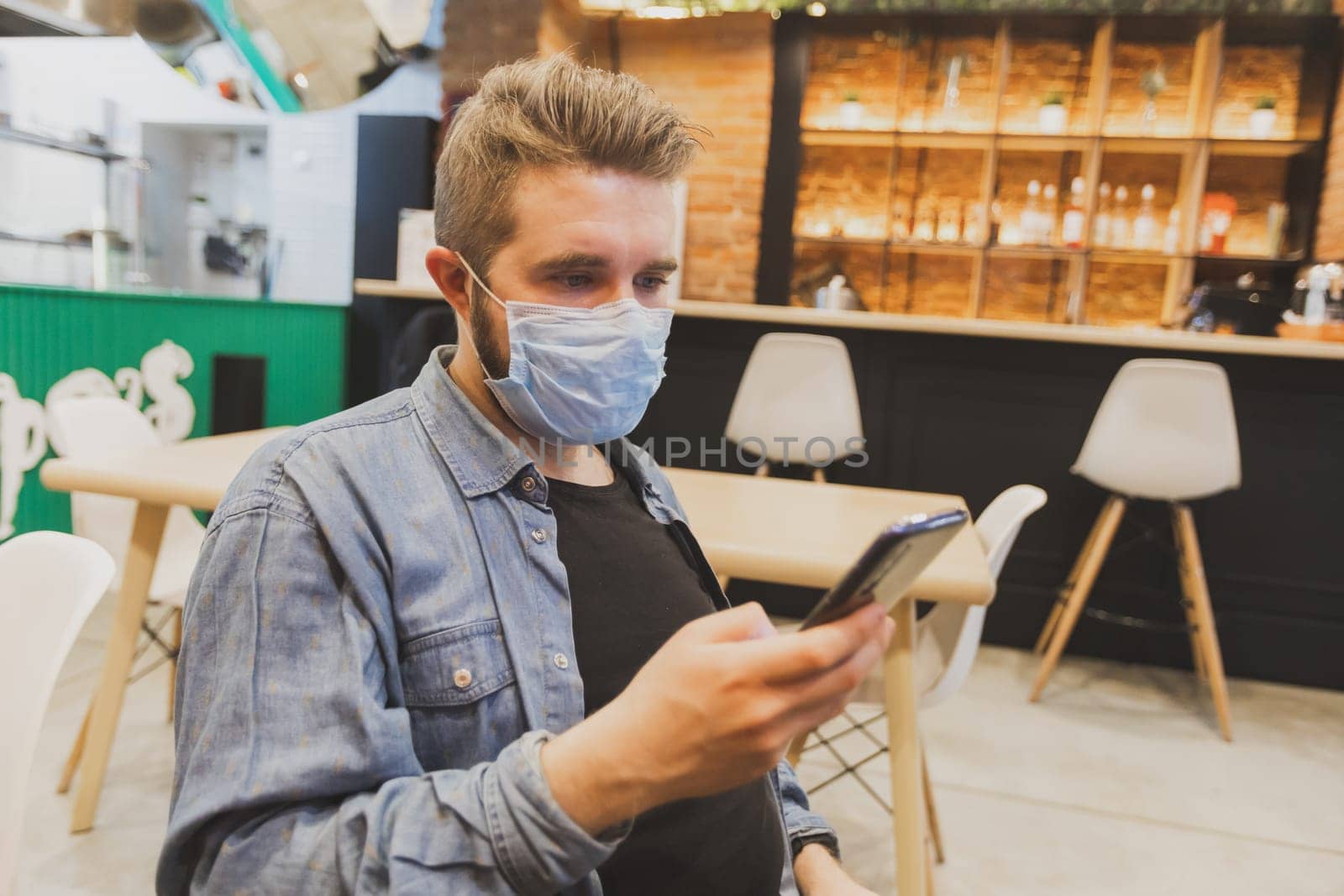 Man wears medical face mask in public area and using smartphone to do work business and social network communication in public cafe work space area. by Satura86