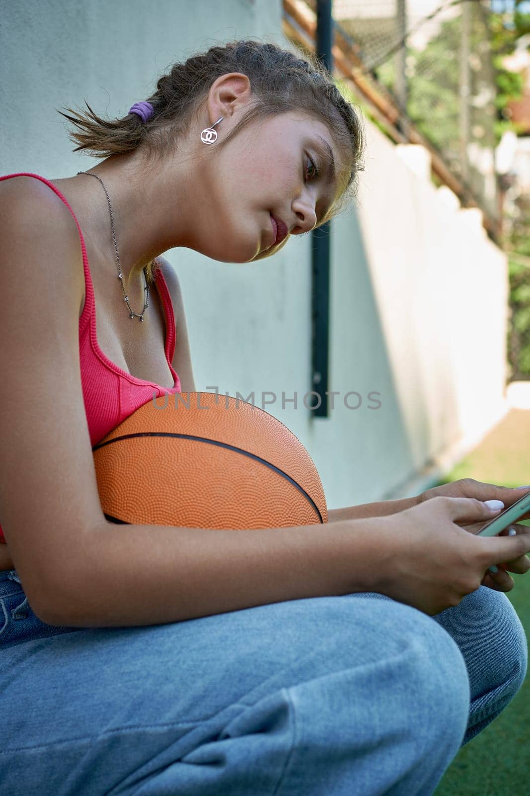 a girl sits with a basketball on the sports ground holding a smartphone in her hands. by AliaksandrFilimonau