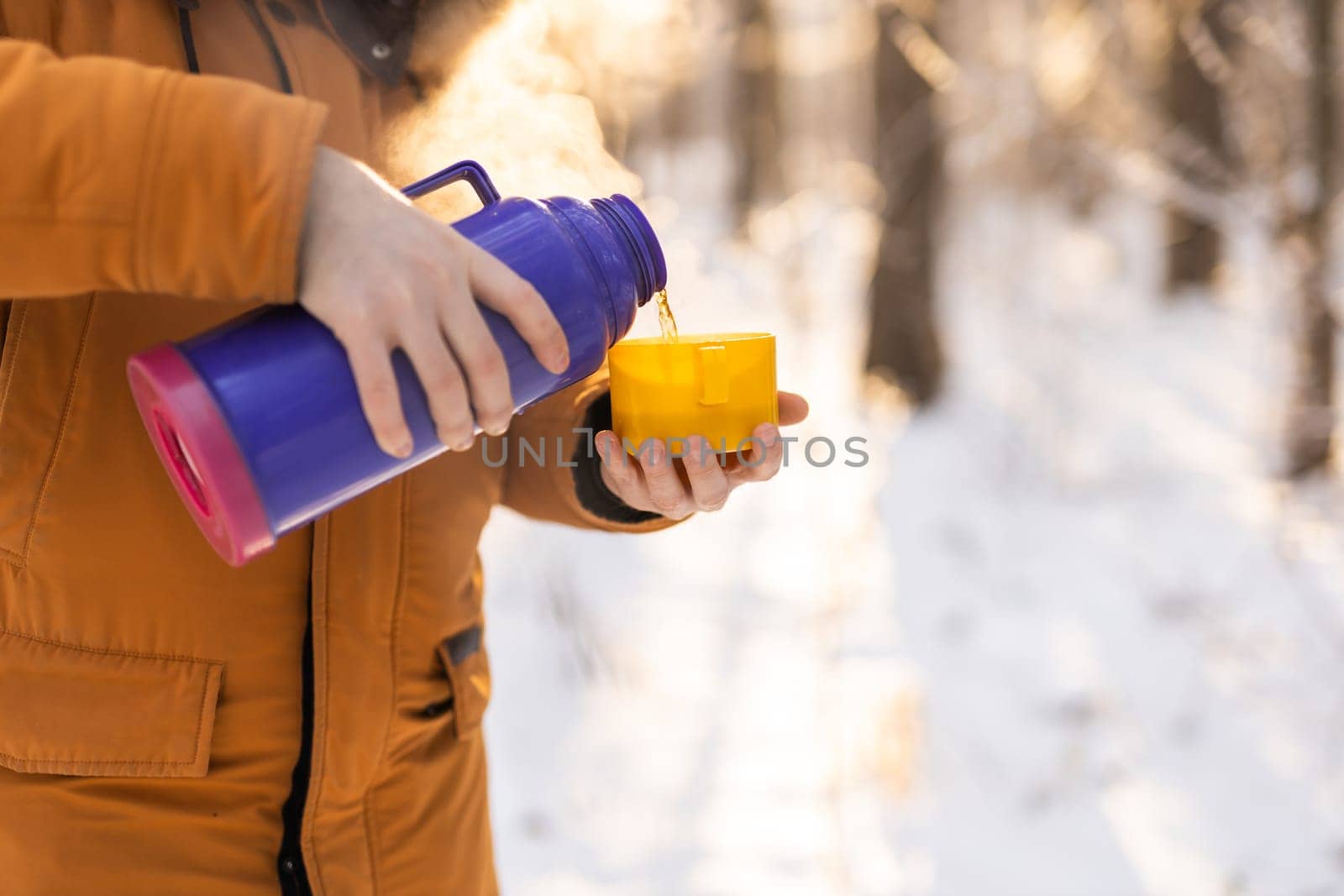 Man pours hot tea from a thermos into a snow walking in snowy frozen winter forest at sunset. Adventure, tourism and camping concept. Copy space and empty place for text advertising.