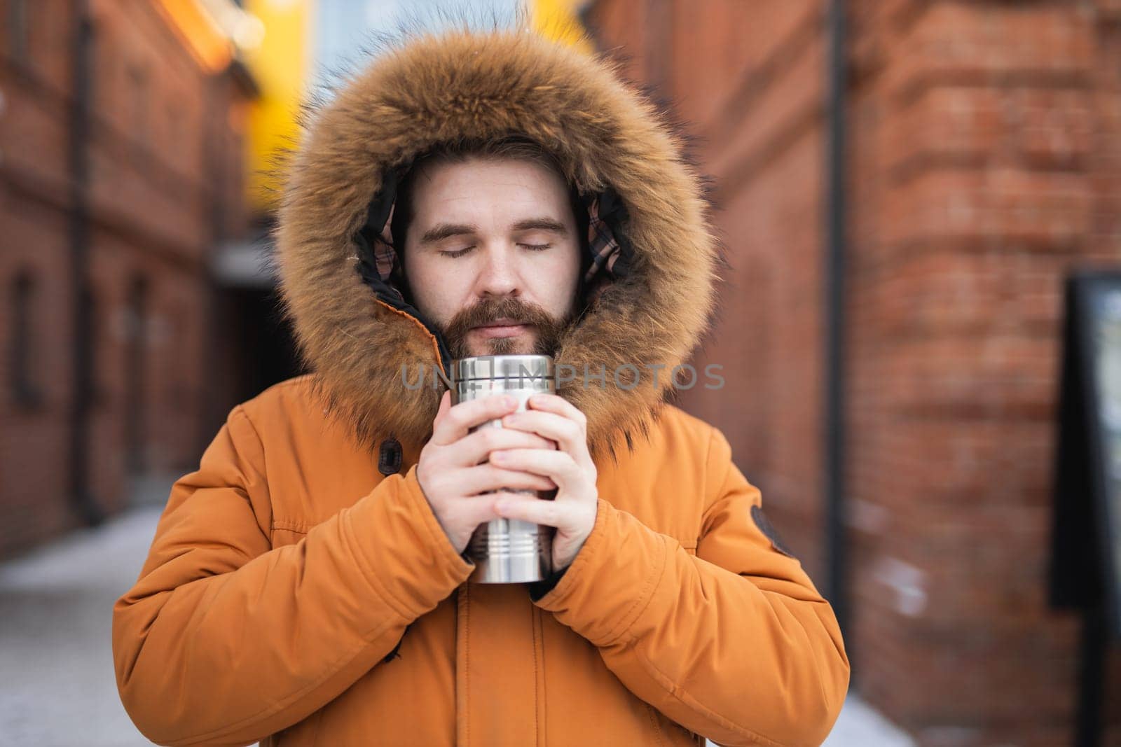 Close-up portrait handsome bearded millennial man in winter clothes and with thermos snow outdoor. Cold season and hot beverage in winter time.