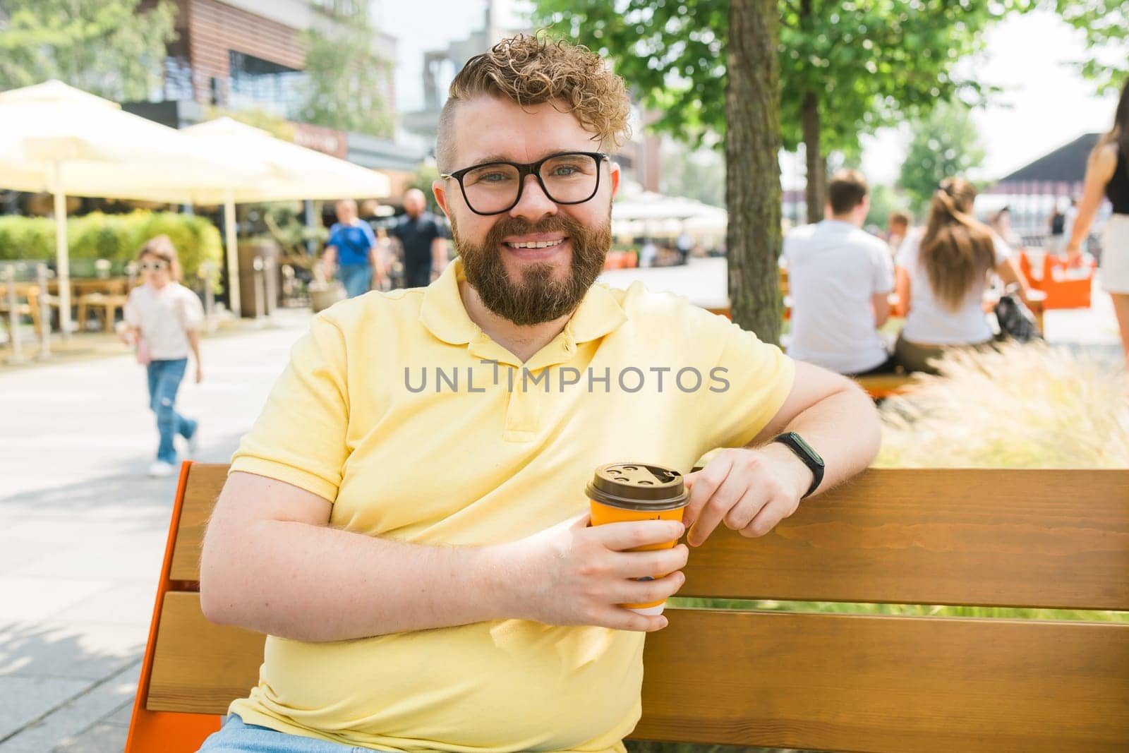Millennial smiling student man in yellow polo t-shirt eyeglasses sitting on bench drink coffee have breakfast rest relax in sunshine spring green city park outdoors on nature. Urban leisure concept
