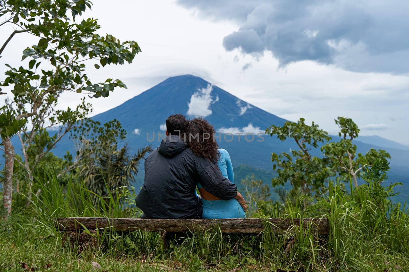 A couple of lovers are sitting on a bench, hugging each other and enjoying the view of the popular sacred cloud-covered Mount Agung on the island of Bali
