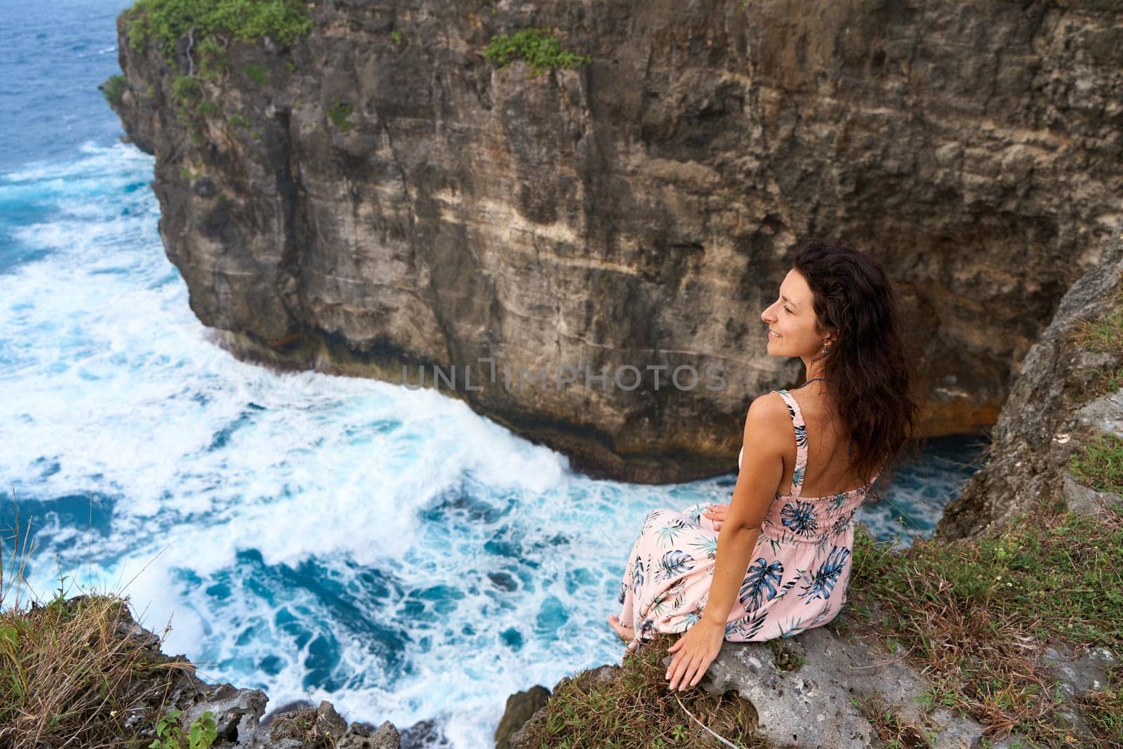 A beautiful woman in a pink dress sits on a cliff above the ocean on the island of Nusa Penida. Devil's Billabong an incredibly wonderful lagoon with splashes from the waves. by Try_my_best