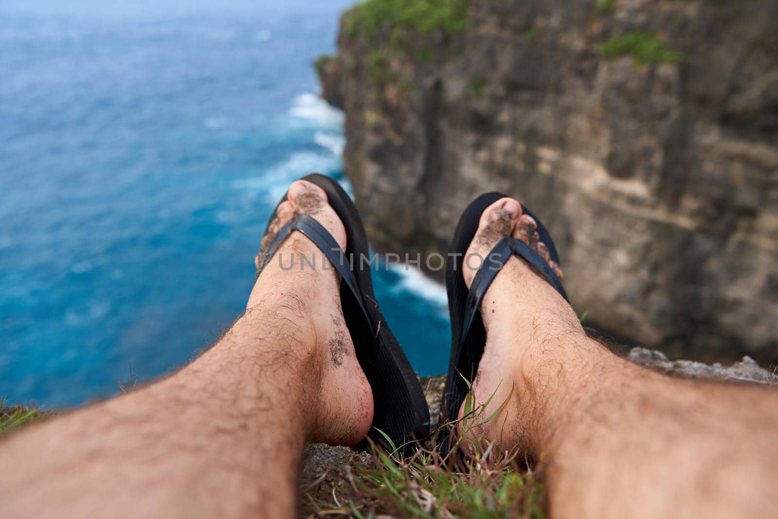 A man sits on a cliff with his legs dangling over an abyss above the ocean. First person photo