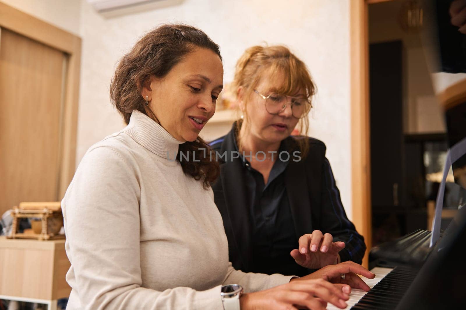 Multi ethnic young adult woman learning playing the pianoforte, sitting near her teacher who explains the truth position of fingers on piano keys during individual music lesson indoor