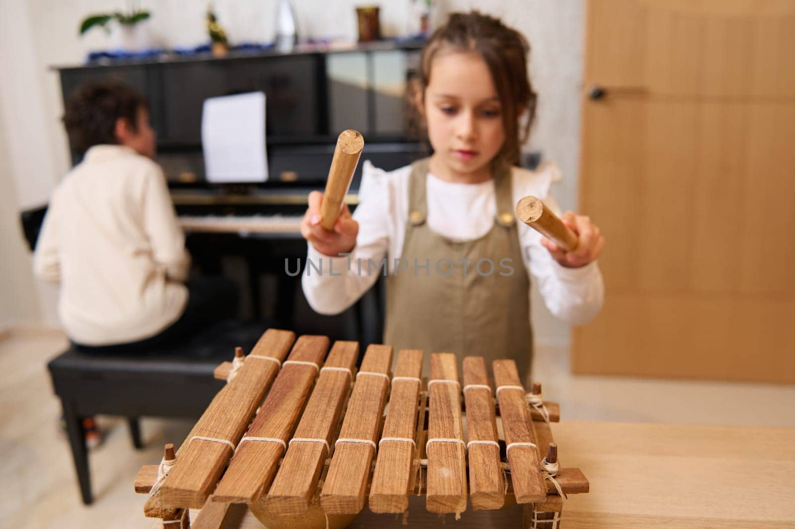 Little Caucasian child girl beating with wooden mallets on xylophone, playing music against her brother sitting at grand piano. Kids education and entertainment. Music lesson. Leisure and hobbies