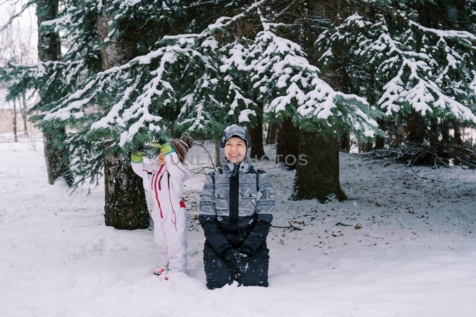 Smiling mother in a ski suit sits on her knees in the snow near a small child standing near a snow-covered pine tree by Nadtochiy