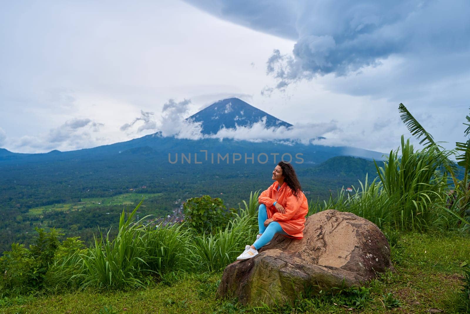 A young woman sits on a large rock in a viewpoint and enjoys the view of the sacred Mount Agung volcano hidden by clouds on a rainy day on the island of Bali.
